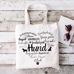 May include: A white canvas tote bag featuring a black graphic design in the shape of a heart. The design includes the German word Hund, various dog-related terms, silhouettes of dogs, and a paw print. The bag has two top handles for carrying.
