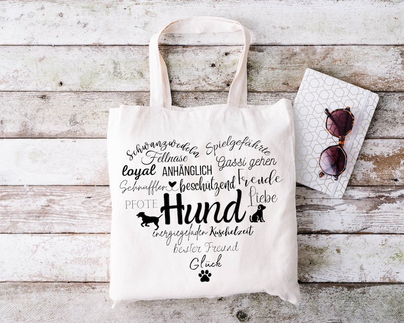 May include: A white canvas tote bag featuring a black graphic design in the shape of a heart. The design includes the German word Hund, various dog-related terms, silhouettes of dogs, and a paw print. The bag has two top handles for carrying.