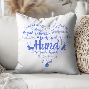 May include: A white square throw pillow featuring a blue heart-shaped graphic composed of German words related to dogs, including the central word Hund, along with silhouettes of a running dog, a sitting dog, and a single paw print at the bottom.
