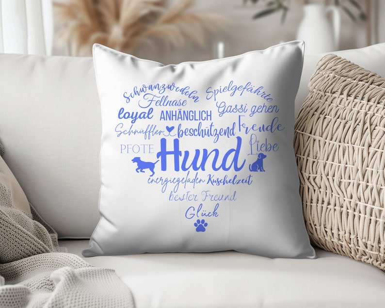 May include: A white square throw pillow featuring a blue heart-shaped graphic composed of German words related to dogs, including the central word Hund, along with silhouettes of a running dog, a sitting dog, and a single paw print at the bottom.