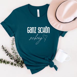 May include: Teal blue t-shirt with white text that reads "Ganz sch&ouml;n schr&auml;g" with a heart symbol.