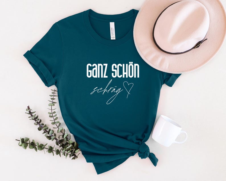 May include: Teal blue t-shirt with white text that reads "Ganz sch&ouml;n schr&auml;g" with a heart symbol.