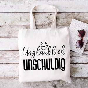May include: A white tote bag with black text that reads "Unglaublich unschuldig" and a smiley face. The bag is sitting on a white wooden surface with a notebook and sunglasses.