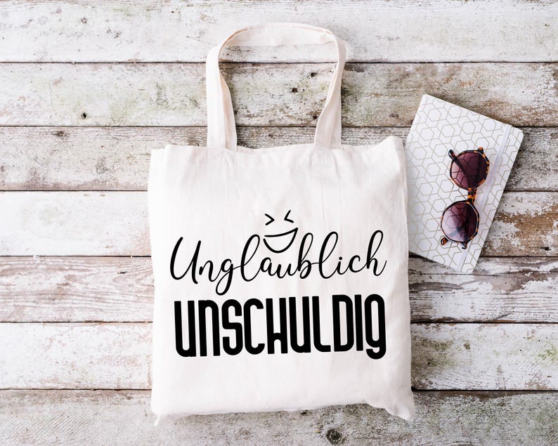May include: A white tote bag with black text that reads "Unglaublich unschuldig" and a smiley face. The bag is sitting on a white wooden surface with a notebook and sunglasses.