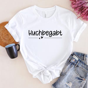 May include: White t-shirt with black text that reads "Huchbegabt" with two small hearts underneath the text.