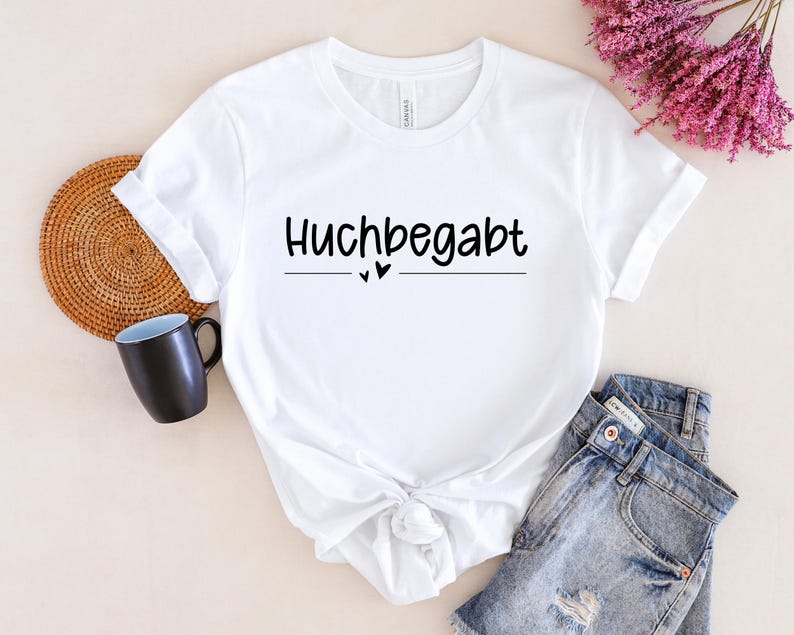 May include: White t-shirt with black text that reads "Huchbegabt" with two small hearts underneath the text.