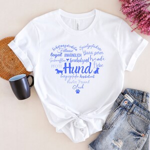 May include: A white short-sleeved t-shirt featuring a blue graphic print in the shape of a heart. The design includes the word Hund, various German words related to dogs, small dog silhouettes, and a paw print centered on the chest area.