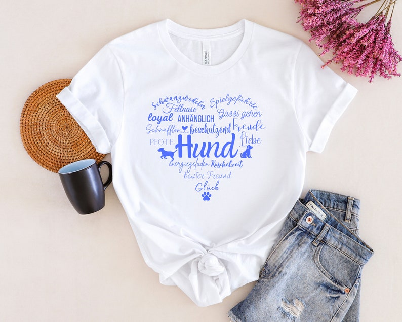 May include: A white short-sleeved t-shirt featuring a blue graphic print in the shape of a heart. The design includes the word Hund, various German words related to dogs, small dog silhouettes, and a paw print centered on the chest area.