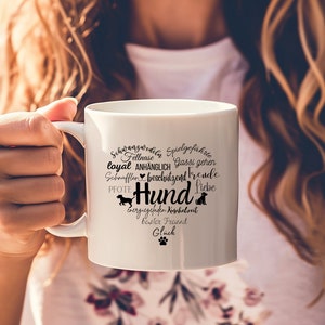 May include: A white ceramic mug featuring a heart-shaped graphic composed of various German words related to dogs, including the central word Hund, along with small silhouettes of dogs and a paw print in black ink on the front surface.
