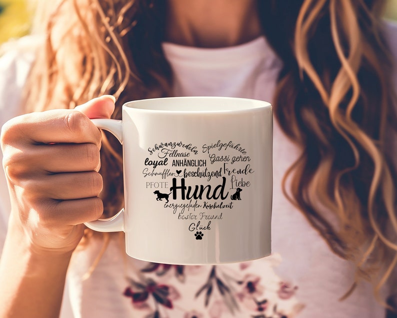 May include: A white ceramic mug featuring a heart-shaped graphic composed of various German words related to dogs, including the central word Hund, along with small silhouettes of dogs and a paw print in black ink on the front surface.