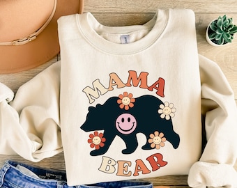 May include: A cream colored sweatshirt with the text "Mama Bear" in a retro font. The text is surrounded by a black bear silhouette with colorful flowers and smiley faces.
