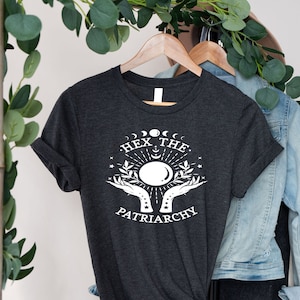 May include: A dark gray t-shirt with a white graphic design featuring a crescent moon, stars, and hands holding a crystal ball. The text "HEX THE PATRIARCHY" is printed below the graphic.