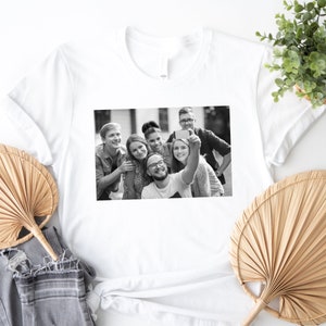 May include: Black and white photo of a group of friends taking a selfie. The friends are smiling and appear to be having fun. The photo is printed on a white t-shirt.