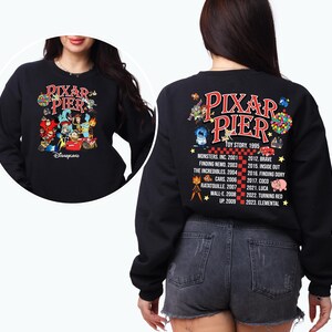 May include: Black sweatshirt with a graphic of Pixar characters on the front and a list of Pixar movies and release dates on the back. The text reads "Pixar Pier" and "Disneyland".