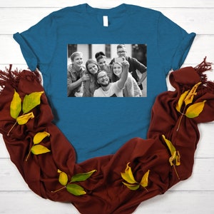 May include: A teal blue t-shirt with a black and white photo of a group of friends taking a selfie. The photo shows six people smiling and looking at the camera.