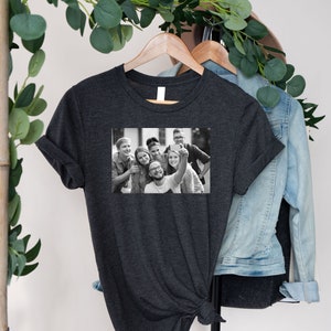 May include: A dark heather grey t-shirt with a black and white photo of a group of friends taking a selfie. The photo shows six people smiling and looking at the camera.