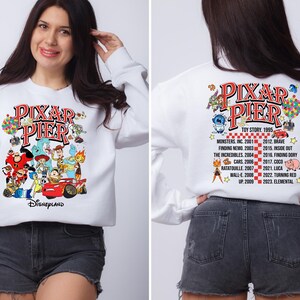 May include: White sweatshirt with a colorful graphic of Pixar characters and the text "Pixar Pier Disneyland". The back of the sweatshirt has a list of Pixar movies and their release years.