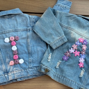 May include: Two light blue denim jackets. One jacket features the letter "I" made of pink, white, and purple yarn flowers. The other jacket has the letter "A" made of pink and purple yarn flowers. Both jackets have distressed details.
