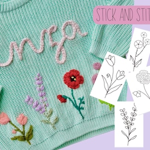 May include: A light green knitted sweater with the word "Enza" embroidered in pink yarn. The sweater also features embroidered flowers in red, pink, purple, and green. The image includes four separate sheets of paper with black line drawings of flowers, labeled "Stick and Stitch".