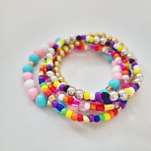 May include: A stack of colorful beaded bracelets. The bracelets feature a variety of colors, including pink, blue, yellow, orange, green, purple, silver, and gold. The beads are small and round, creating a vibrant and playful accessory.