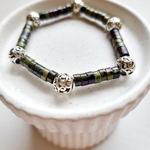 May include: A green and black beaded bracelet with silver charm beads. The bracelet is on a white surface.
