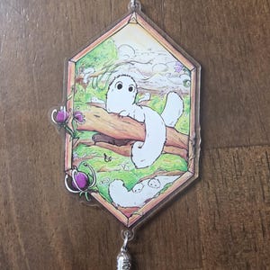 May include: A clear acrylic keychain featuring a whimsical illustration of a white creature with large eyes resting on a log. The keychain has a blue tassel and a silver-colored metal clasp. The background shows a forest scene with purple flowers.