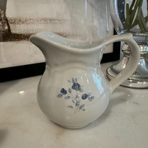 Könnte beinhalten: Ein kleiner, weißer Keramikkrug mit rundem Körper und gebogenem Henkel. Der Krug hat ein blaues Blumendesign auf der Vorderseite. Der Ausguss ist sanft geschwungen. Der Krug wird wahrscheinlich zum Servieren von Flüssigkeiten verwendet.