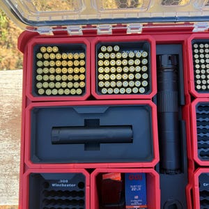 May include: A red and black organizer case with a clear lid, filled with ammunition and gun accessories. The case contains various compartments holding brass cartridges, a black cylindrical object, and a box of CCI .22 LR ammunition.