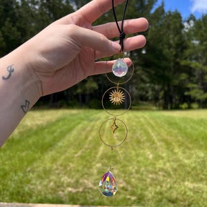 May include: A gold sun and star charm with crystal teardrop prisms hanging from a black string. The charms are suspended from a gold ring.