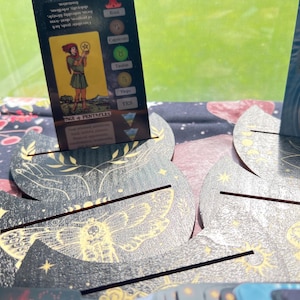 May include: A tarot card featuring an illustration of a person holding a pentacle, with text that reads "Page of Pentacles." The card is propped up in a decorative card holder with a moth design. The background is a blurred green.