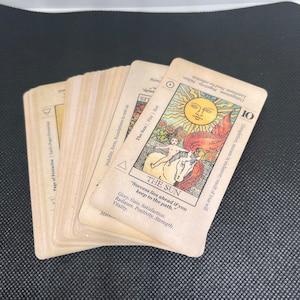 May include: A deck of tarot cards with a vintage aesthetic. The cards are beige with black text and illustrations. One card features a sun illustration and the text "THE SUN". The deck is fanned out on a dark surface.
