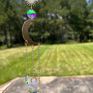 May include: A gold crescent moon suncatcher with iridescent crystals and a clear crystal teardrop hanging from it. The suncatcher is hanging in front of a green grassy field.