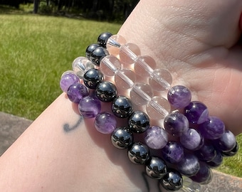 May include: A stack of three bracelets made with amethyst, clear quartz, and hematite beads. The bracelets are worn on a person's wrist.