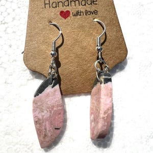 May include: A pair of silver dangle earrings with pink and black stone drops. The earrings are handmade and have a tag that says "Handmade with love".
