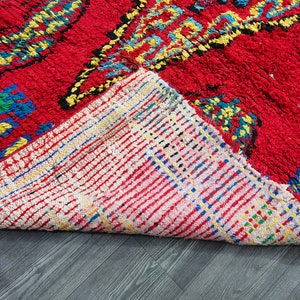 May include: Close-up of the back side of a red, blue, and yellow patterned rug. The rug is folded over, revealing a white, pink, and blue woven backing.