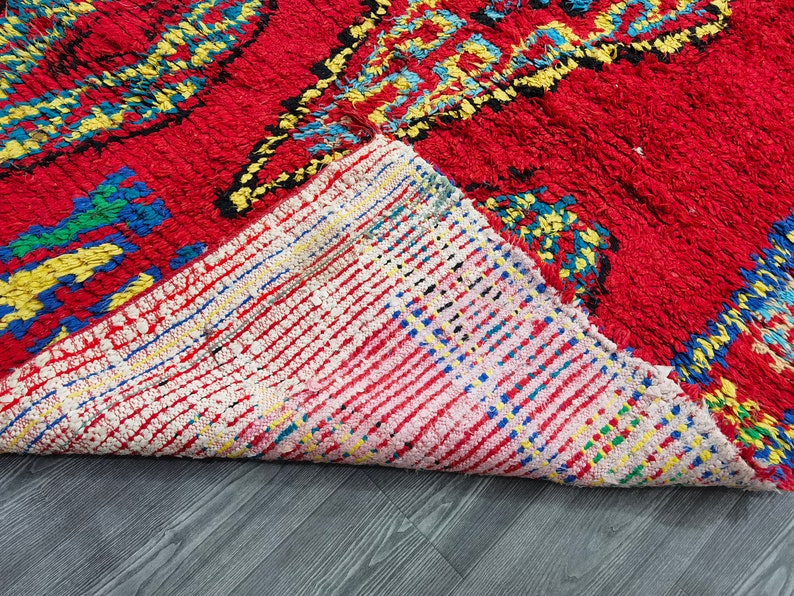 May include: Close-up of the back side of a red, blue, and yellow patterned rug. The rug is folded over, revealing a white, pink, and blue woven backing.