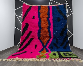 May include: A vibrant, hand-woven rug with a bold, abstract design. The rug features a striking combination of hot pink, navy blue, and orange hues, accented with black geometric shapes. The rug has a fringed edge and is displayed against a black and white wall.