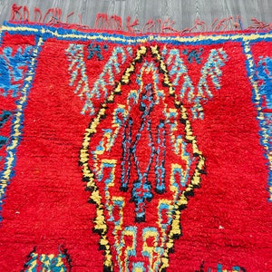 May include: A red Moroccan rug with a blue, yellow, and black geometric pattern. The rug has a fringe border.