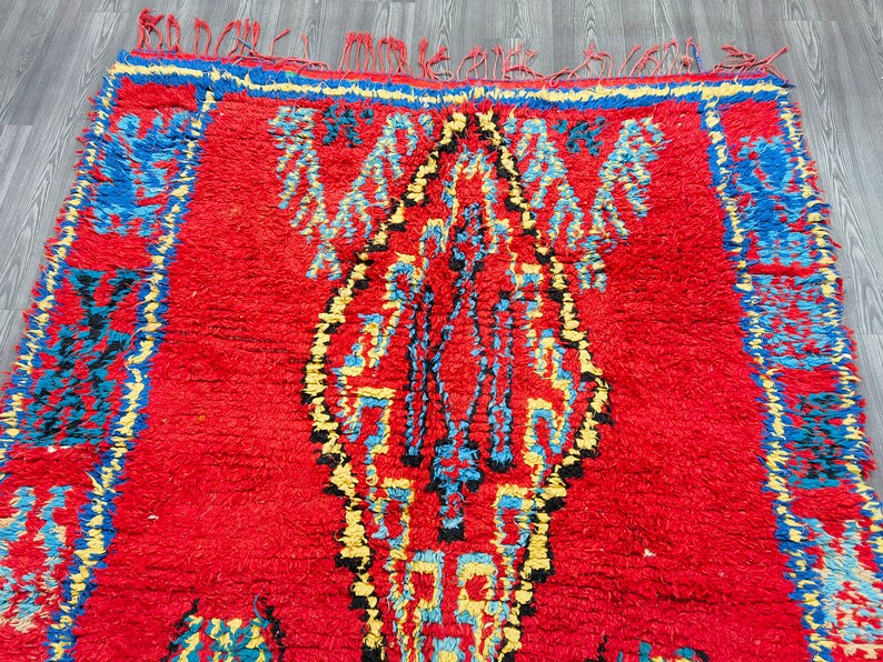 May include: A red Moroccan rug with a blue, yellow, and black geometric pattern. The rug has a fringe border.