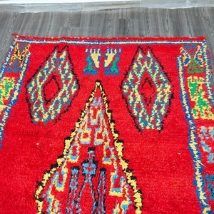 May include: A red Moroccan rug with a geometric pattern in shades of blue, yellow, and green. The rug has a thick, fluffy pile and is woven with intricate details.