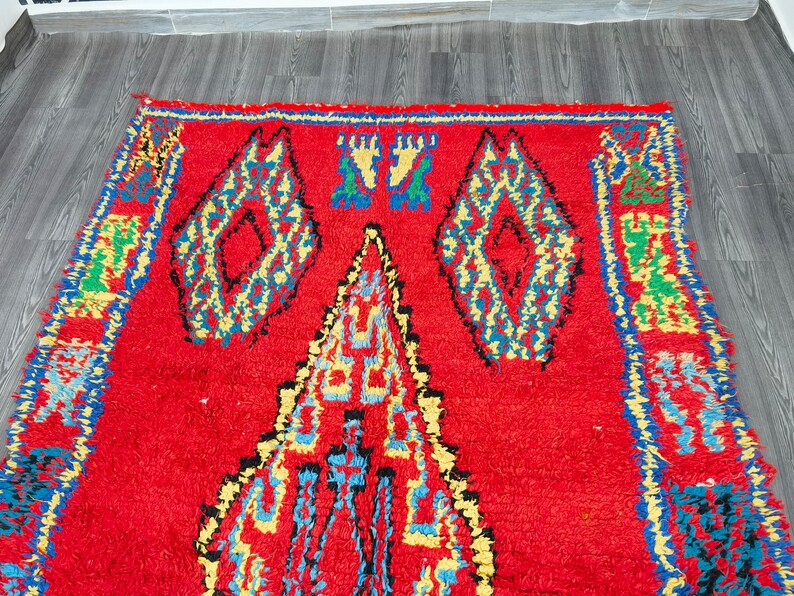 May include: A red Moroccan rug with a geometric pattern in shades of blue, yellow, and green. The rug has a thick, fluffy pile and is woven with intricate details.