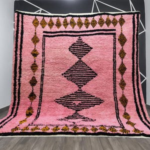 May include: Pink Moroccan rug with black and gold geometric patterns. The rug features a central diamond design and borders with repeating diamond shapes. The rug is displayed on a wooden floor.