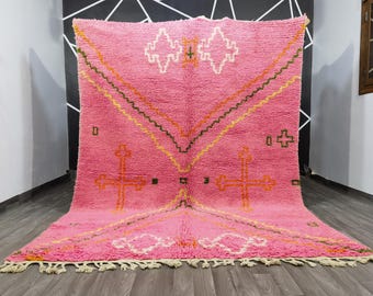 May include: A vibrant pink rug with a geometric design. The rug features a central cross-like pattern in orange, with white, yellow, and green accents. The edges are finished with a fringe.