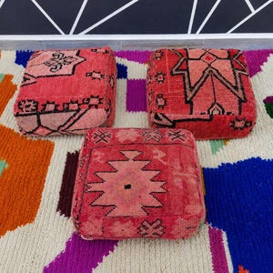May include: Three square floor cushions in shades of red and pink, each featuring distinct black geometric patterns. The cushions are arranged on a multi-colored rug with large abstract shapes in orange, white, blue, purple, and green.