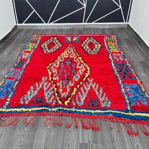 May include: A red Moroccan rug with a traditional geometric pattern in shades of blue, yellow, and black. The rug has a fringe border and is made of wool.