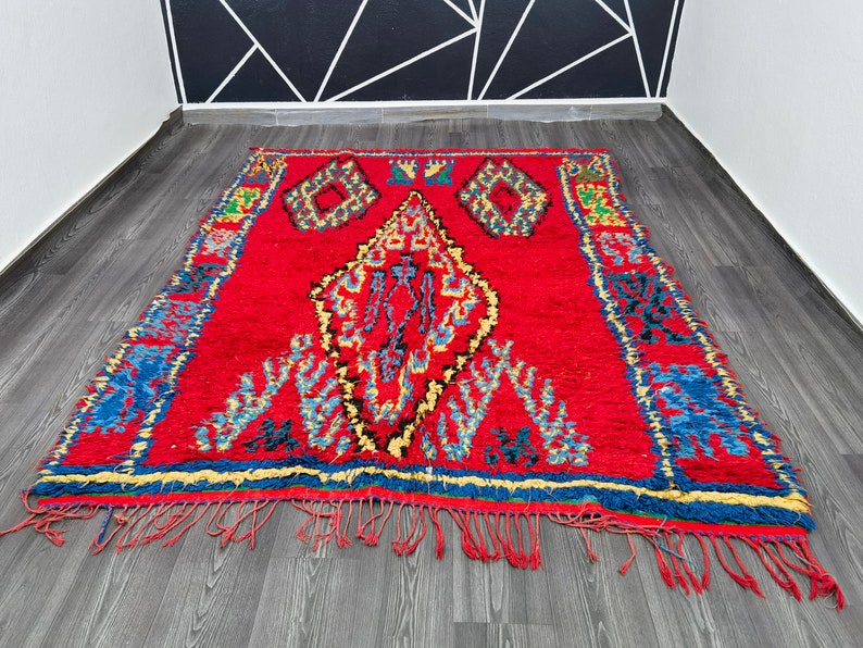 May include: A red Moroccan rug with a traditional geometric pattern in shades of blue, yellow, and black. The rug has a fringe border and is made of wool.