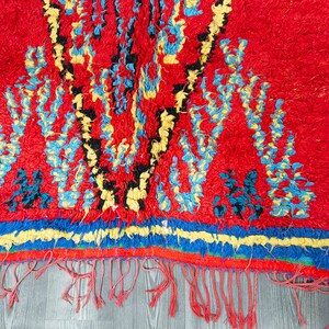 May include: A red, blue, yellow, and black Moroccan rug with a geometric pattern. The rug has a fringe border and is made of wool.