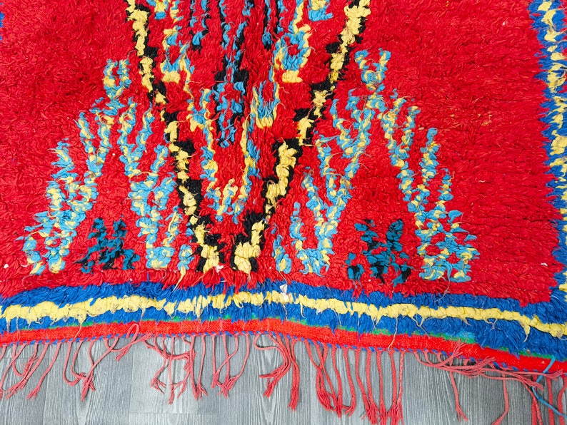 May include: A red, blue, yellow, and black Moroccan rug with a geometric pattern. The rug has a fringe border and is made of wool.