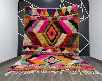 Puede incluir: Una colorida alfombra marroquí hecha a mano con un diseño geométrico. La alfombra presenta un patrón de diamantes central, rodeado de rayas y triángulos en tonos rosa, amarillo, verde y azul. La alfombra tiene flecos en el borde inferior.