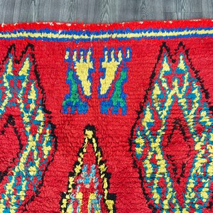 May include: A red, hand-woven rug with a geometric pattern in yellow, blue, and green. The rug has a fringe border.